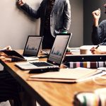 A group of people in a business meeting sit around a table with laptops showing graphs while one person stands at the whiteboard, pointing to it.